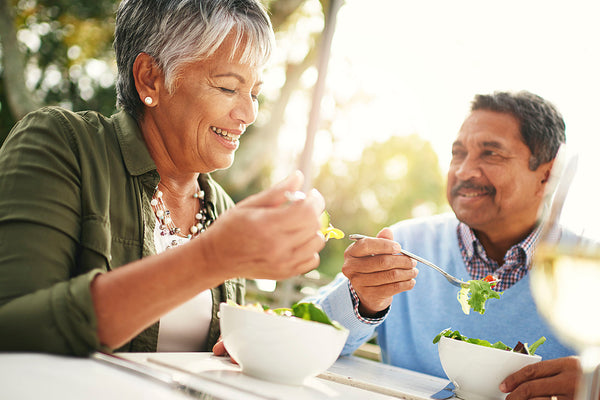 Older man and woman eating salads out of white bowls together outside in the sunshine