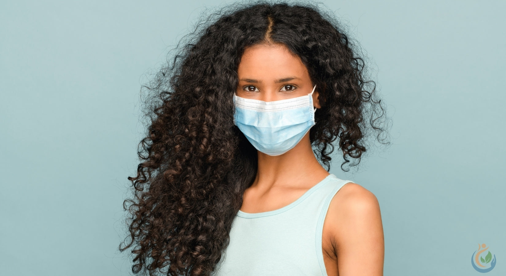 Beautiful woman with long curly, dark hair and with a covid-19 face mask on a light blue background