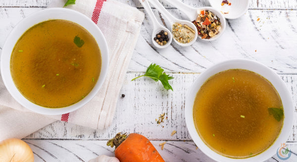 two bowls of bone broth with various vegetables and spices on white wooden table